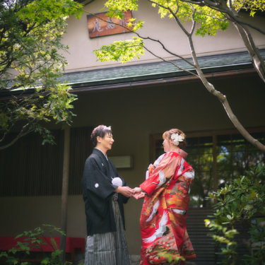 新緑のもみじが茂る日本庭園の茶室前で、両手を取り合って見つめ合う赤い色打掛の花嫁と黒紋付袴の新郎。和装前撮りの一枚