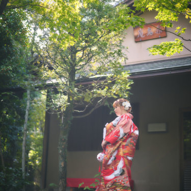 緑の木々に囲まれた茶室の前で、鶴柄の赤い色打掛の後ろ姿を見せて振り返る花嫁。和装の柄が映える前撮り