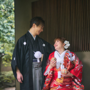 格子の壁の軒下で、鞠の小物を手に新郎と笑い合う色打掛の花嫁。白い胡蝶蘭の髪飾りが華やかな和装の結婚写真
