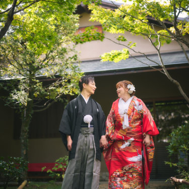 新緑のもみじの下、石畳を手をつないで歩く新郎新婦。赤い色打掛と黒紋付袴のコントラストが美しい和装前撮り