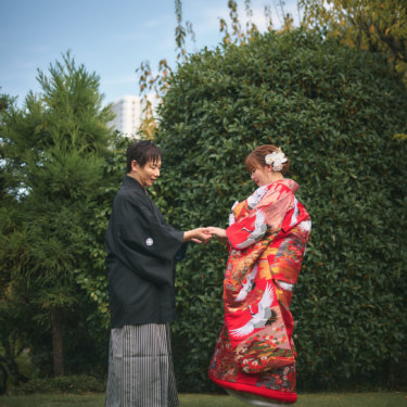 芝生の庭で手を取り合って向かい合う、黒紋付袴の新郎と鶴柄の赤い色打掛の花嫁。青空の下の和装ロケーションフォト
