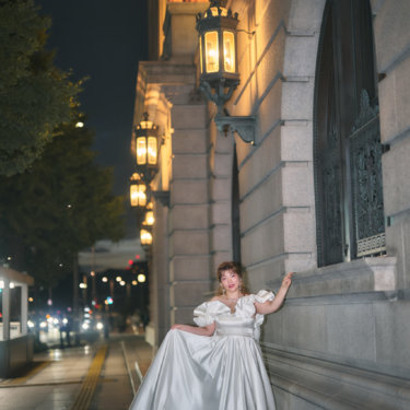 ランプが灯る石造りの建物の前で、サテンのドレスの裾を持ってポーズをとる花嫁。夜の丸の内で撮るクラシカルな前撮り