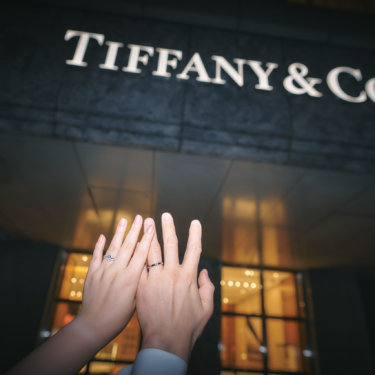 夜の店舗のファサードを背に、指輪をはめた手を重ねて掲げる新郎新婦の手元。婚約指輪と結婚指輪を写すウェディングフォト