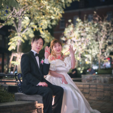 ライトアップされた木々の下のベンチで、赤い糸を小指に結んで笑う新郎新婦。小物を使ったかわいい夜の前撮り
