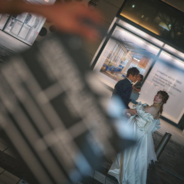 手前のカチンコをぼかし、ショーウィンドウの前で抱き合う新郎新婦を写した映画風の結婚写真。丸の内の夜の街並みで撮影