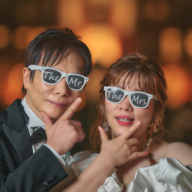 玉ボケの夜景を背に、文字入りの白いサングラスをかけてポーズを決める新郎新婦。おちゃめな小物使いの前撮り