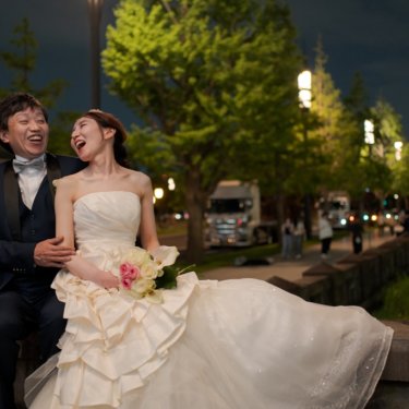 ライトアップされた並木道沿いの石垣に並んで座り、大笑いする新郎とフリルのドレスの花嫁の夜の前撮り