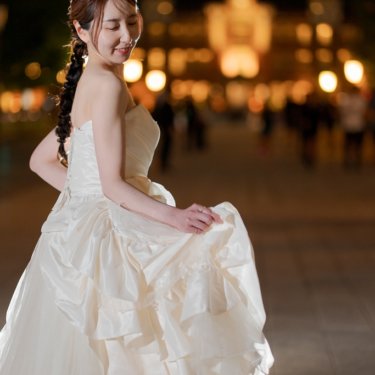 街の夜景をぼかした背景で、フリルのドレスの裾をつまんで振り返るティアラの花嫁のソロカット。夜の前撮り