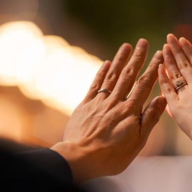 夜の街の光をぼかした背景で、結婚指輪をはめた手を重ねるように合わせる新郎新婦の手元カット。前撮りの指輪ショット