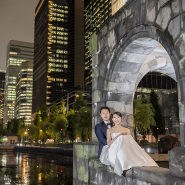 お濠の水面にビルの明かりが映る夜、石造りのアーチの上に並んで座る新郎新婦。丸の内の夜景フォトウェディング
