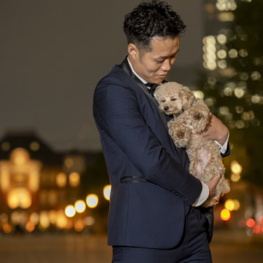 街の灯りを背に、愛犬のトイプードルを胸に抱いて優しく見つめるタキシードの新郎のソロカット。愛犬と前撮り