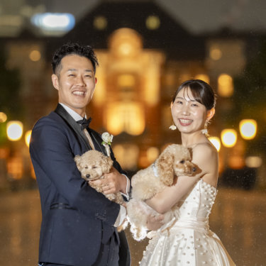 夜の駅前で愛犬を抱き、カメラに笑顔を向ける新郎とウェディングドレスの花嫁。家族で残す結婚写真