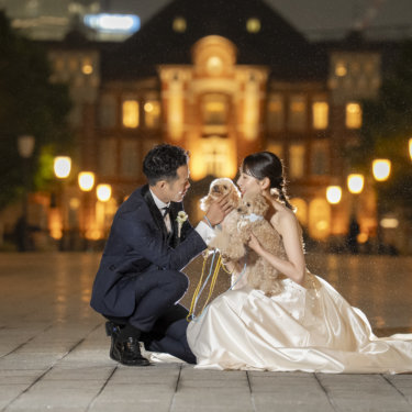 ライトアップされた赤レンガ駅舎の前でしゃがみ込み、愛犬に顔を寄せる新郎新婦。愛犬と一緒の前撮り