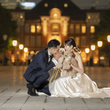 夜の駅前広場で愛犬をはさんで顔を寄せ合う新郎とウェディングドレスの花嫁。温かな雰囲気のフォトウェディング