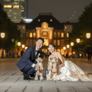 東京駅の夜景を背に、しゃがんで2匹の愛犬と並び笑顔を向ける新郎新婦。ペットと一緒の結婚写真