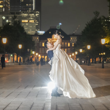 夜の東京駅を背に、背後からのライトで輝くウェディングドレスの花嫁を抱き上げる新郎の前撮り