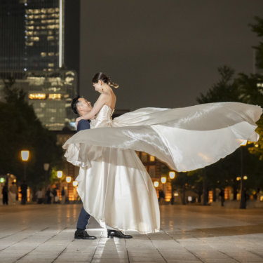 街灯が並ぶ夜の駅前広場で、花嫁を抱き上げてドレスの裾を大きくなびかせる新郎。躍動感のある結婚写真