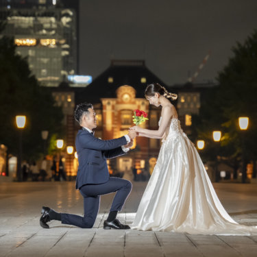 夜の東京駅前広場でひざまずき、赤いバラを渡す新郎と受け取るウェディングドレスの花嫁のフォトウェディング
