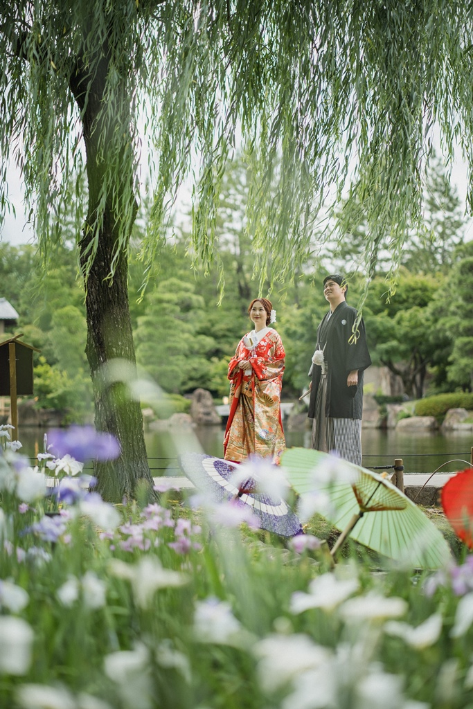 柳の枝越し、池のほとりで朱色の色打掛の新婦と黒紋付の新郎が寄り添う和装ロケーションフォト。