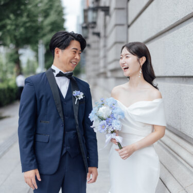 石造りの壁沿いで笑い合うネイビータキシードの新郎と、ブルーの紫陽花ブーケを持つオフショルダードレスの花嫁の結婚写真