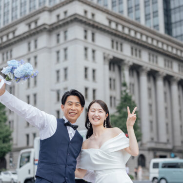 丸の内の重厚な石造りのビルを背に、ブーケを掲げる新郎とピースする花嫁。笑顔あふれる洋装の前撮り写真