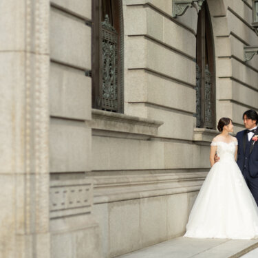 装飾窓と重厚な扉が並ぶクラシカルな石造りの建物の前で、寄り添う新郎新婦を写した前撮り