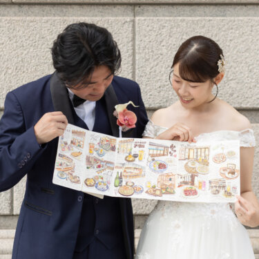 石の壁の前でイラストマップをのぞき込む新郎新婦。小物でデートのようなストーリーを演出した洋装前撮り