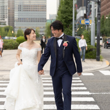 手をつないで笑いながら横断歩道を歩くタキシードの新郎とウェディングドレスの花嫁。東京駅前の広場で撮る自然体の結婚写真