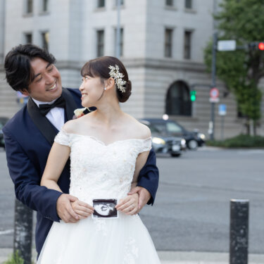 丸の内の交差点で、後ろから花嫁を抱き寄せてエコー写真を見せる新郎。ほほえましいマタニティ前撮りの一枚