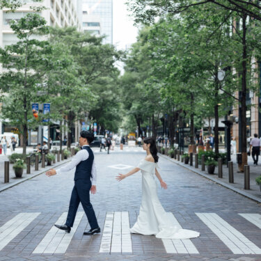石畳の丸の内仲通りの横断歩道で、両手を広げて向かい合う新郎新婦。並木道の奥行きを活かした映画風の前撮り
