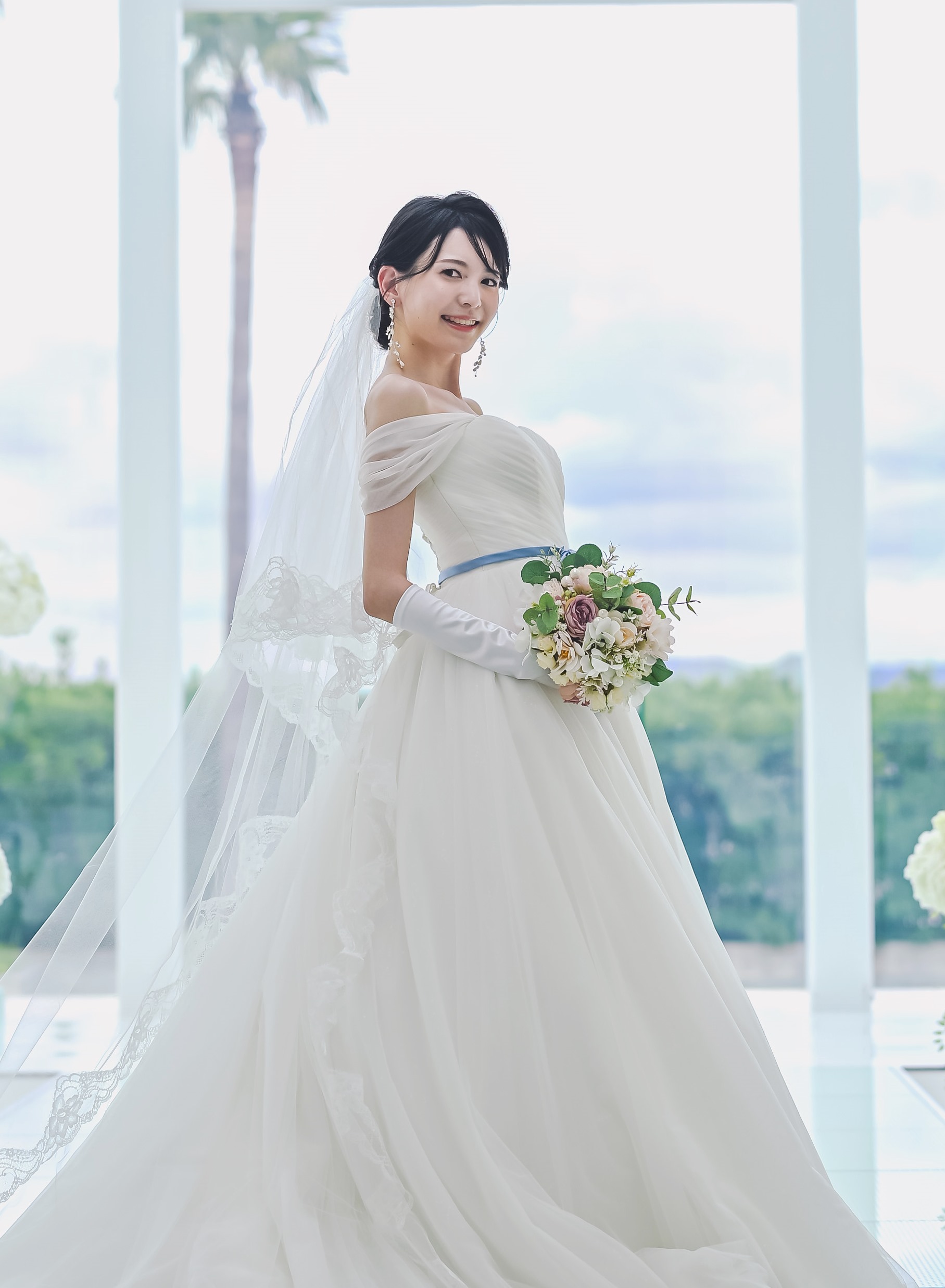 ブルーのリボンベルトが映えるオフショルダーのウェディングドレスを着た新婦、ヤシの木を背景に。