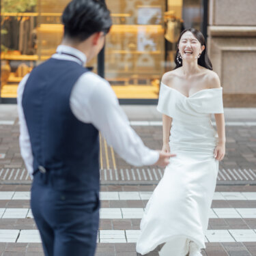横断歩道の向こうで笑顔を見せるマーメイドドレスの花嫁と、手を差し伸べる新郎の後ろ姿を捉えたウェディングフォト