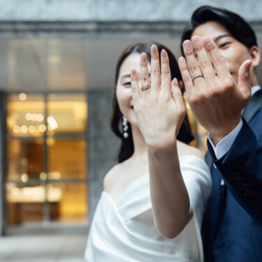 新郎新婦がそろって手の甲をカメラに向け、結婚指輪を見せる前撮り写真。ショーウィンドウのあかりが柔らかくボケている