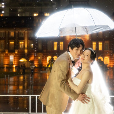 透明のビニール傘を差して頬を寄せ合う新郎新婦。雨に煙る東京駅丸の内駅舎の夜景をバックにした結婚写真