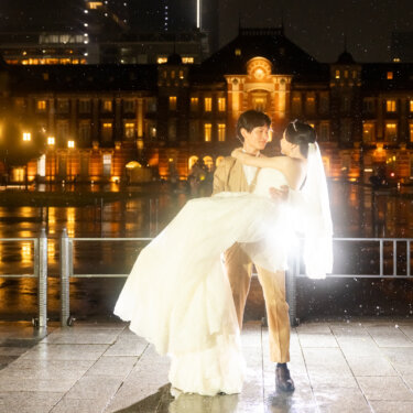 ライトアップされた東京駅丸の内駅舎を背に、新郎が新婦をお姫様抱っこ。雨に濡れた駅前広場が光を映す夜の前撮り