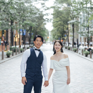 石畳と街路樹が続く丸の内仲通りで、手をつないで並んで立つベスト姿の新郎とウェディングドレスの花嫁の前撮り写真