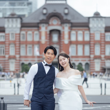 赤レンガの東京駅を背景に、柵に寄りかかって寄り添う蝶ネクタイの新郎とオフショルダードレスの花嫁の洋装フォトウェディング
