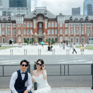 丸の内駅舎と高層ビルを背に、サングラスをかけてしゃがむ新郎新婦。東京駅の前撮りで人気のクールなポーズ