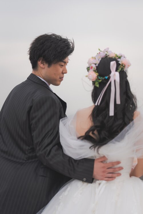 花冠とリボンの髪飾りを付けた花嫁の腰に手を添えて見つめる新郎。ブラックのストライプタキシードで撮るフォトウェディングの後ろ姿カット（ブラックラージ／衣装番号TP08）