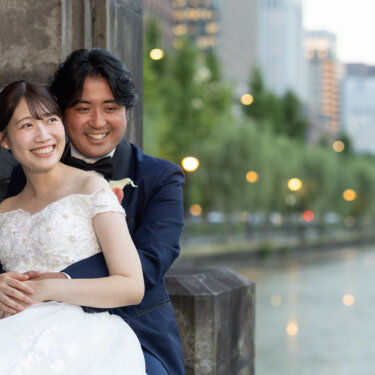 石の壁の前で花嫁を後ろから抱きしめる新郎。お濠と並木をぼかした背景で笑い合う東京の前撮り
