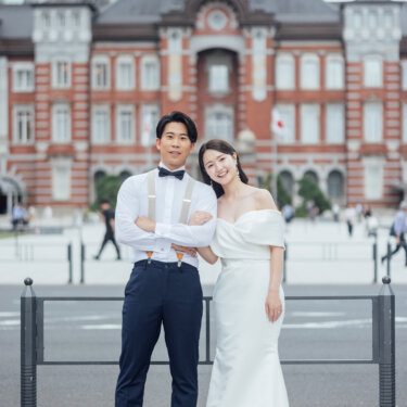 東京駅丸の内駅舎の正面で、サスペンダー姿で腕を組む新郎とマーメイドラインのウェディングドレスの花嫁が並ぶ、王道のフォトウェディング