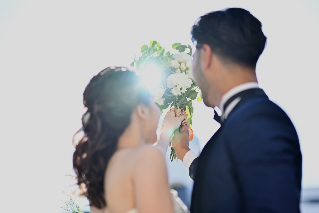 逆光の中、白いブーケを手渡す新郎と受け取る新婦の後ろ姿。