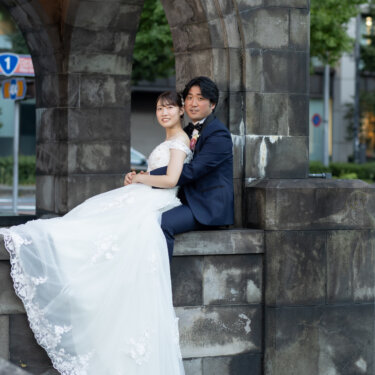 お濠を望む石造りのアーチに腰かけ、レースのトレーンを垂らして寄り添う新郎新婦のウェディングフォト
