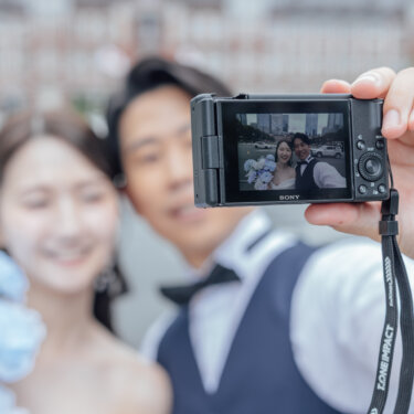 新郎がコンパクトカメラを掲げてふたりで自撮りする瞬間を撮った一枚。液晶に映る笑顔とブルーのブーケがかわいい東京駅での前撮り