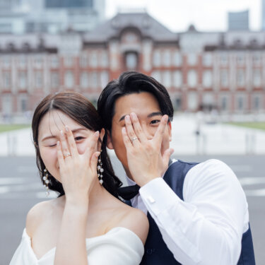 東京駅丸の内駅舎の前で、片手で目元を隠しながら笑う新郎新婦。指輪がのぞく顔隠しポーズで撮るウェディングフォト