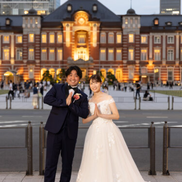 東京駅の駅舎を背に、胸の前で手を合わせて並ぶ新郎とウェディングドレスの花嫁。おそろいのポーズがかわいい前撮り