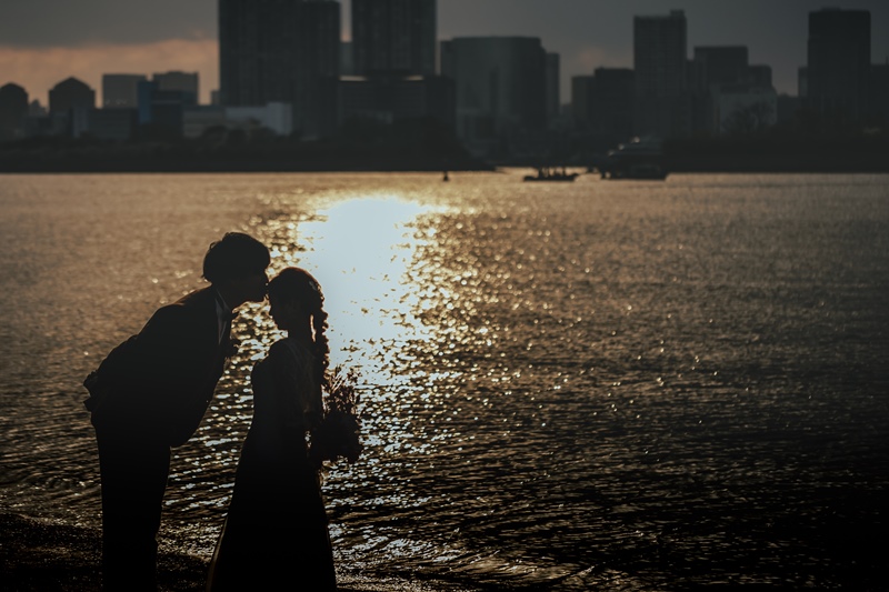 水辺の逆光シルエットで額にキスをする新郎新婦、東京の高層ビル群を背景に。