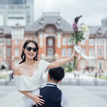 赤レンガの東京駅丸の内駅舎を背に、サングラス姿の花嫁を抱き上げる新郎。花嫁がブーケを掲げる元気いっぱいの洋装前撮り
