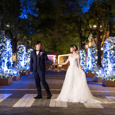 青いイルミネーションの並木道で、新郎が花嫁の手を引いて向き合う夜のワンシーン。東京駅周辺で撮るナイトフォトの前撮り