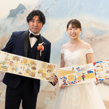 手描きのイラストマップを両手で広げ、並んで笑顔を見せる新郎新婦。マーブル模様の壁を背景に小物で遊び心を出した洋装前撮り